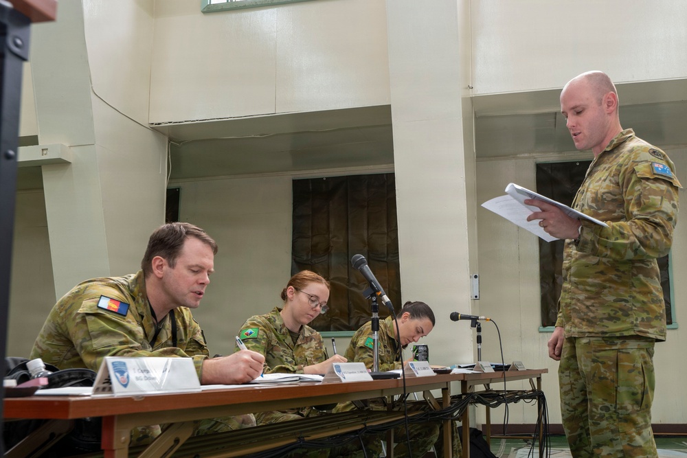 Four Australian Defence Force personnel are engaged in what appears to be a military briefing or formal meeting. Three personnel are seated at a table with name placards and microphones, taking notes, while a fourth standing personnel reads from documents. The setting appears to be an indoor military facility or conference room.
