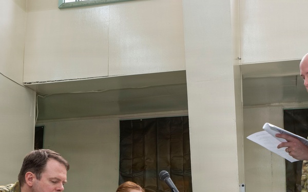 Four Australian Defence Force personnel are engaged in what appears to be a military briefing or formal meeting. Three personnel are seated at a table with name placards and microphones, taking notes, while a fourth standing personnel reads from documents. The setting appears to be an indoor military facility or conference room.