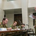 Four Australian Defence Force personnel are engaged in what appears to be a military briefing or formal meeting. Three personnel are seated at a table with name placards and microphones, taking notes, while a fourth standing personnel reads from documents. The setting appears to be an indoor military facility or conference room.