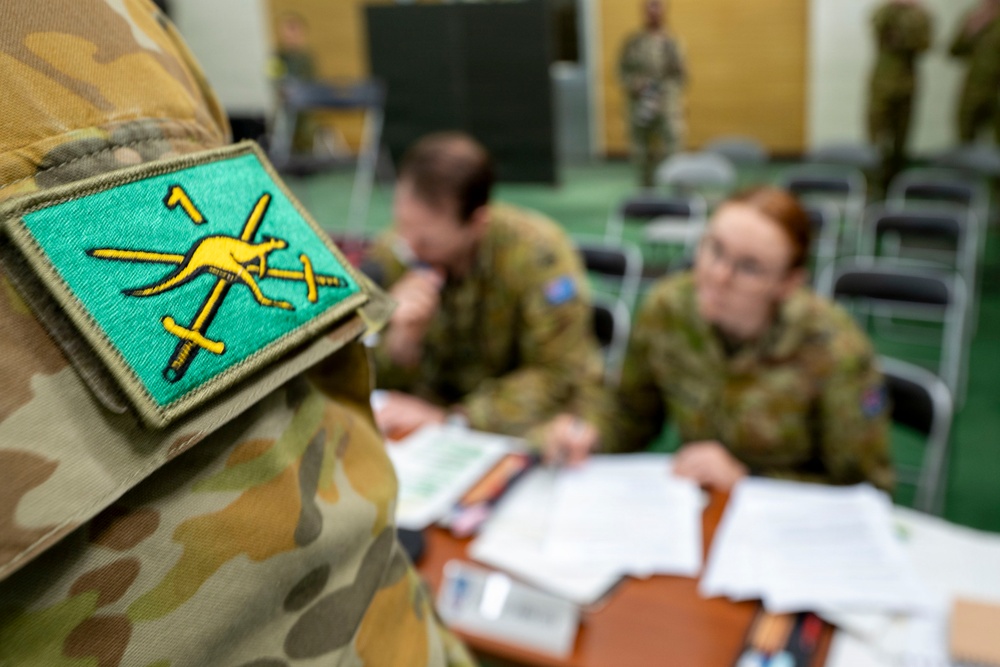 The image shows Australian Army personnel in a briefing or administrative setting, with a close-up focus on a unit patch on a camouflage uniform sleeve. The patch features a yellow kangaroo and crossed swords on a teal/green background, consistent with Australian Army insignia. In the background, soldiers are seated at a table reviewing documents in what appears to be a meeting room.