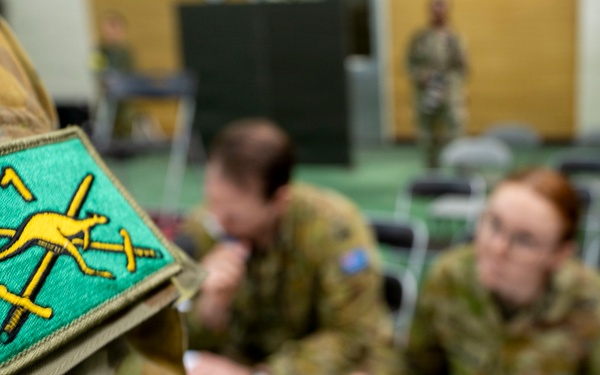 The image shows Australian Army personnel in a briefing or administrative setting, with a close-up focus on a unit patch on a camouflage uniform sleeve. The patch features a yellow kangaroo and crossed swords on a teal/green background, consistent with Australian Army insignia. In the background, soldiers are seated at a table reviewing documents in what appears to be a meeting room.