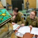 The image shows Australian Army personnel in a briefing or administrative setting, with a close-up focus on a unit patch on a camouflage uniform sleeve. The patch features a yellow kangaroo and crossed swords on a teal/green background, consistent with Australian Army insignia. In the background, soldiers are seated at a table reviewing documents in what appears to be a meeting room.