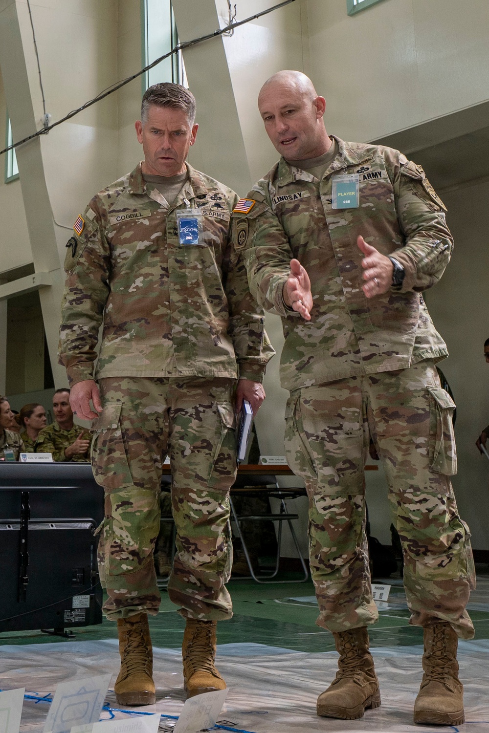Two U.S. Army soldiers in MultiCam/OCP camouflage uniforms are engaged in conversation during what appears to be a military exercise or war game event. The soldier on the right, identified by his name tape as 'LINDSAY,' is gesturing and speaking to the soldier on the left, identified as 'COGBILL.' Both wear credential badges and are standing on what appears to be a large floor map used for operational planning.
