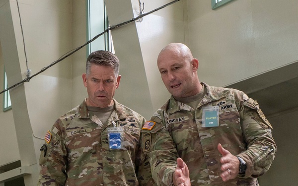 Two U.S. Army soldiers in MultiCam/OCP camouflage uniforms are engaged in conversation during what appears to be a military exercise or war game event. The soldier on the right, identified by his name tape as 'LINDSAY,' is gesturing and speaking to the soldier on the left, identified as 'COGBILL.' Both wear credential badges and are standing on what appears to be a large floor map used for operational planning.