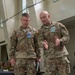 Two U.S. Army soldiers in MultiCam/OCP camouflage uniforms are engaged in conversation during what appears to be a military exercise or war game event. The soldier on the right, identified by his name tape as 'LINDSAY,' is gesturing and speaking to the soldier on the left, identified as 'COGBILL.' Both wear credential badges and are standing on what appears to be a large floor map used for operational planning.