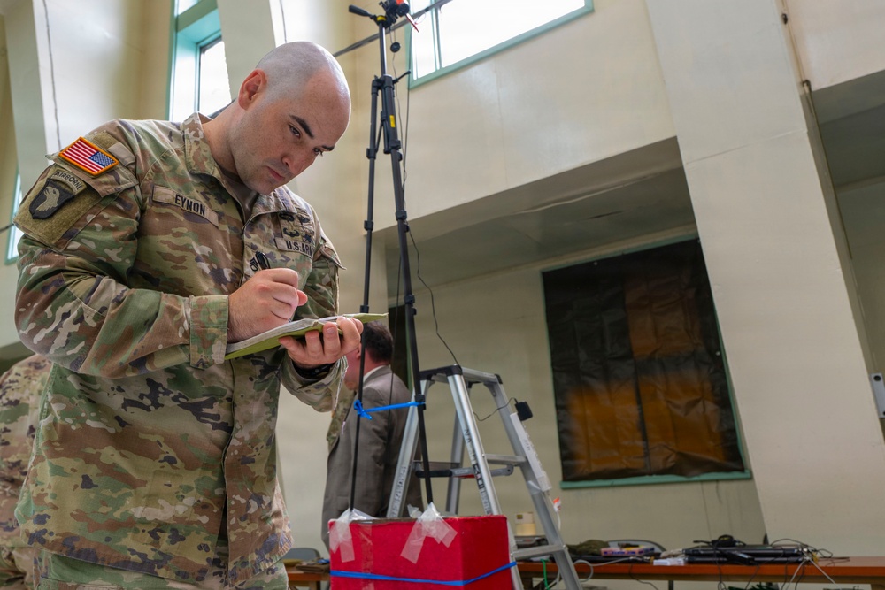 A U.S. Army soldier identified by name tape as 'EYNON' is writing notes on a small notepad in what appears to be an indoor facility. He is wearing OCP (Operational Camouflage Pattern) uniform with an American flag patch and airborne tab on the left shoulder, and U.S. Army tape on the chest. In the background, a civilian is working near a ladder and tripod-mounted equipment.