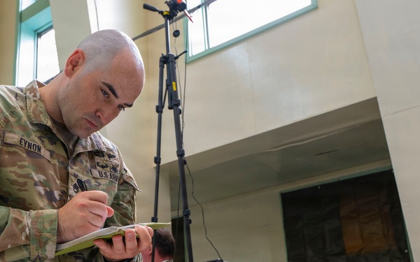A U.S. Army soldier identified by name tape as 'EYNON' is writing notes on a small notepad in what appears to be an indoor facility. He is wearing OCP (Operational Camouflage Pattern) uniform with an American flag patch and airborne tab on the left shoulder, and U.S. Army tape on the chest. In the background, a civilian is working near a ladder and tripod-mounted equipment.