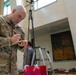 A U.S. Army soldier identified by name tape as 'EYNON' is writing notes on a small notepad in what appears to be an indoor facility. He is wearing OCP (Operational Camouflage Pattern) uniform with an American flag patch and airborne tab on the left shoulder, and U.S. Army tape on the chest. In the background, a civilian is working near a ladder and tripod-mounted equipment.