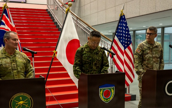 A military press conference is being held at Camp Asaka, Japan, for exercise Yama Sakura 91 (YS-91). Three military officers — appearing to represent Australia, Japan, and the United States — stand at separate podiums in front of their respective national flags. A banner in English and Japanese identifies the event as the YS-91 Press Conference at Camp Asaka.