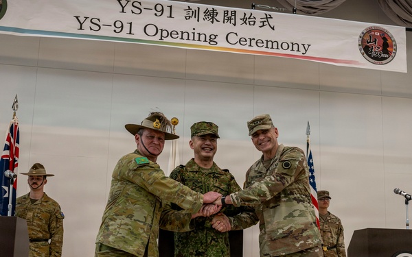 This image depicts the YS-91 Opening Ceremony, a multinational military exercise held at Camp Asaka, Japan. Three military officers representing Australia, Japan, and the United States are shaking hands together in a show of trilateral cooperation. Flags and honor guards from the participating nations are visible in the background.