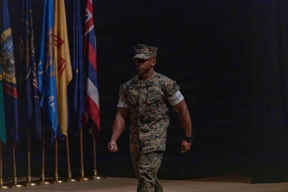 A U.S. Marine in woodland MARPAT camouflage uniform is walking across what appears to be a stage or ceremonial hall. Multiple U.S. state flags are visible on stands in the background to the left. The setting suggests a formal military ceremony or official event.