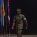 A U.S. Marine in woodland MARPAT camouflage uniform is walking across what appears to be a stage or ceremonial hall. Multiple U.S. state flags are visible on stands in the background to the left. The setting suggests a formal military ceremony or official event.