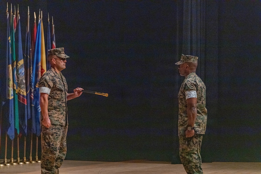Two U.S. Marine Corps personnel in MARPAT digital camouflage uniforms face each other during what appears to be a formal military ceremony. The Marine on the left is holding a ceremonial baton or staff, suggesting this is likely a relief and appointment or change of responsibility ceremony. A row of military and state flags is visible in the background on a darkened stage.