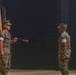 Two U.S. Marine Corps personnel in MARPAT digital camouflage uniforms face each other during what appears to be a formal military ceremony. The Marine on the left is holding a ceremonial baton or staff, suggesting this is likely a relief and appointment or change of responsibility ceremony. A row of military and state flags is visible in the background on a darkened stage.