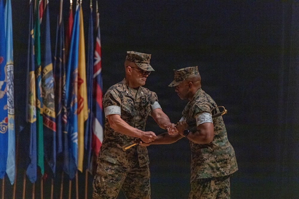 Two U.S. Marine Corps officers in MARPAT camouflage uniforms are engaged in a formal military ceremony, appearing to exchange or pass a ceremonial sword or baton. The background features a row of military and organizational flags, suggesting this is a change of command or similar official military ceremony. The setting appears to be an indoor venue with dark staging.
