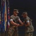 Two U.S. Marine Corps officers in MARPAT camouflage uniforms are engaged in a formal military ceremony, appearing to exchange or pass a ceremonial sword or baton. The background features a row of military and organizational flags, suggesting this is a change of command or similar official military ceremony. The setting appears to be an indoor venue with dark staging.