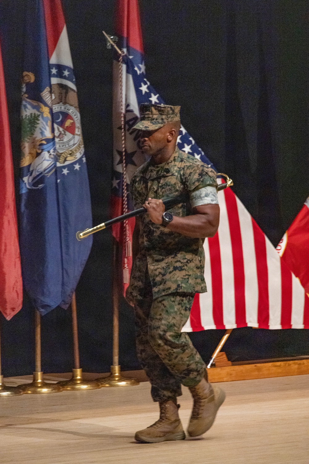 A U.S. Marine in woodland MARPAT camouflage uniform is carrying a ceremonial sword (NCO sword) while marching across a wooden stage floor. Multiple flags are displayed on stands in the background, including the U.S. flag, what appears to be the Missouri state flag, and other military or state flags. The setting appears to be a formal military ceremony held indoors.
