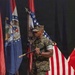 A U.S. Marine in woodland MARPAT camouflage uniform is carrying a ceremonial sword (NCO sword) while marching across a wooden stage floor. Multiple flags are displayed on stands in the background, including the U.S. flag, what appears to be the Missouri state flag, and other military or state flags. The setting appears to be a formal military ceremony held indoors.
