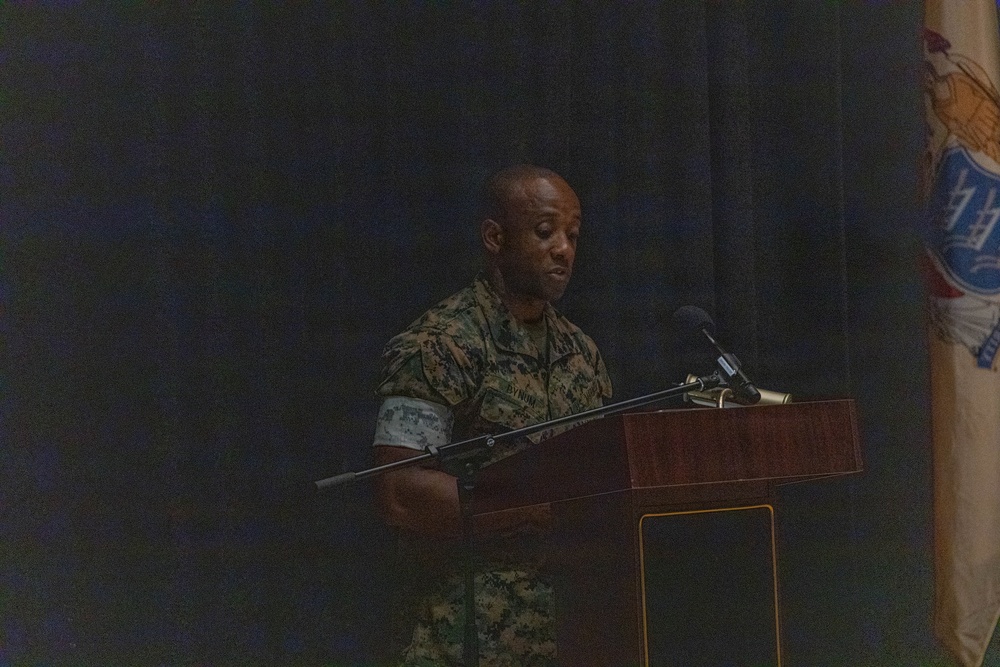 A military service member in camouflage uniform stands at a wooden podium, appearing to deliver a speech or address. The setting appears to be a formal military ceremony or briefing held indoors on a darkened stage. A partially visible flag or banner is present on the right side of the image.
