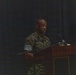 A military service member in camouflage uniform stands at a wooden podium, appearing to deliver a speech or address. The setting appears to be a formal military ceremony or briefing held indoors on a darkened stage. A partially visible flag or banner is present on the right side of the image.