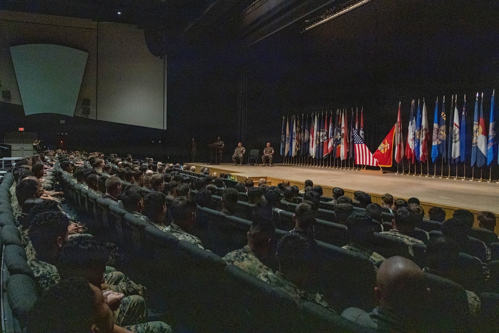 A large military auditorium or theater filled with uniformed personnel seated in rows facing a stage. On the stage, a speaker stands at a podium while two other individuals are seated nearby, with a prominent display of multiple military unit and organizational flags arranged in a row behind them. The event appears to be a formal military briefing, ceremony, or all-hands meeting.