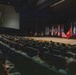 A large military auditorium or theater filled with uniformed personnel seated in rows facing a stage. On the stage, a speaker stands at a podium while two other individuals are seated nearby, with a prominent display of multiple military unit and organizational flags arranged in a row behind them. The event appears to be a formal military briefing, ceremony, or all-hands meeting.