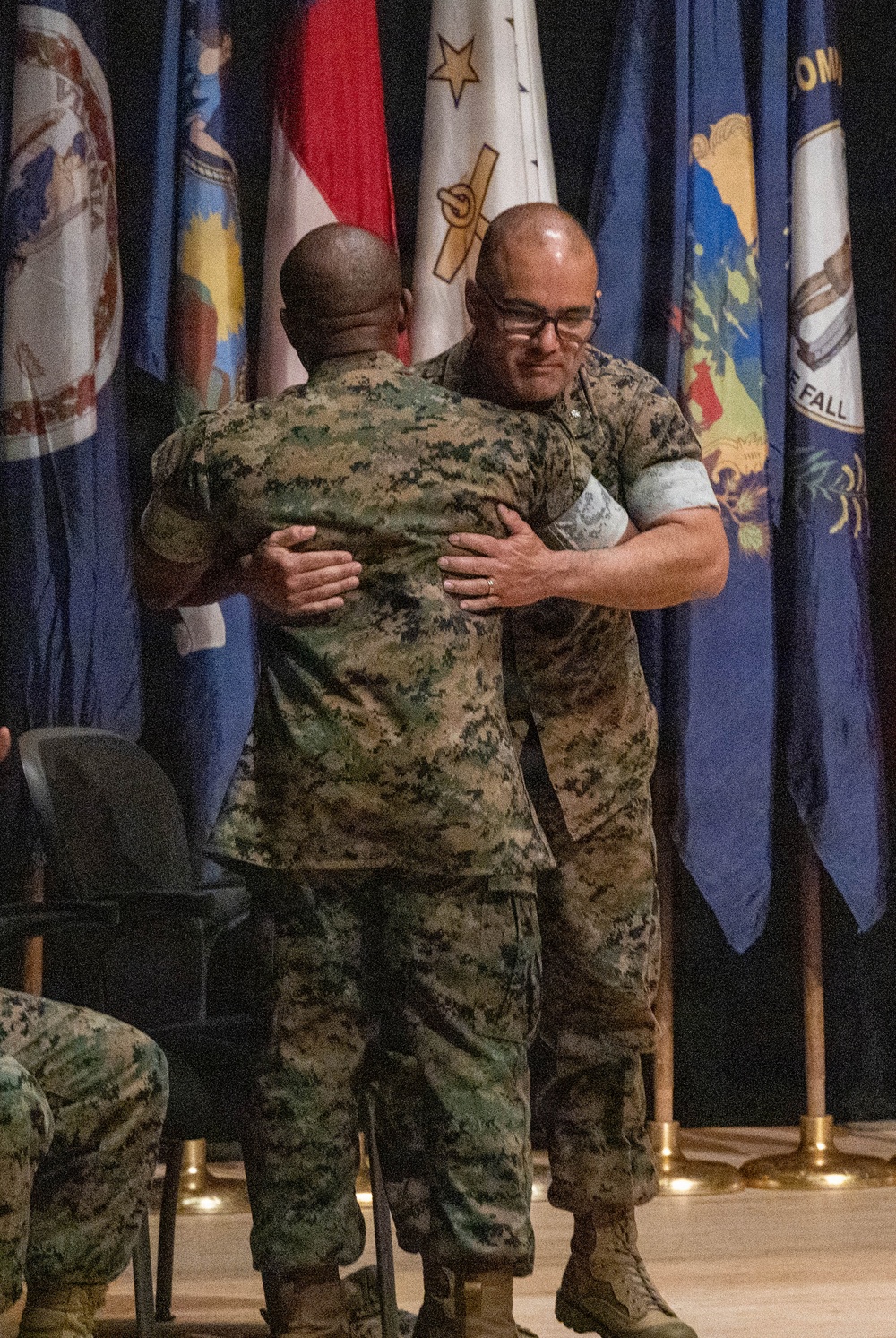Two military personnel in Marine Corps MARPAT camouflage uniforms are embracing on a stage during what appears to be a formal ceremony. Multiple flags are displayed in the background, including what appear to be state flags and a military unit flag. The setting suggests a change of command, retirement, or awards ceremony.