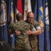 Two military personnel in Marine Corps MARPAT camouflage uniforms are embracing on a stage during what appears to be a formal ceremony. Multiple flags are displayed in the background, including what appear to be state flags and a military unit flag. The setting suggests a change of command, retirement, or awards ceremony.