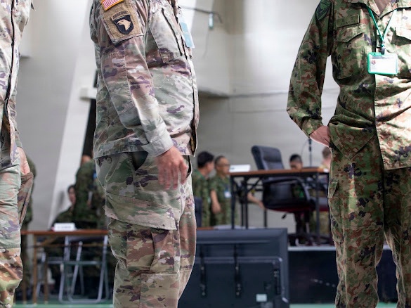 The image shows military personnel from at least two different nations standing in what appears to be a command or operations center. A U.S. Army soldier wearing OCP (Operational Camouflage Pattern) with a 101st Airborne Division patch and American flag is visible alongside personnel in a different camouflage pattern consistent with Japan Ground Self-Defense Force (JGSDF) uniforms. The setting suggests a joint military exercise or coordination meeting.