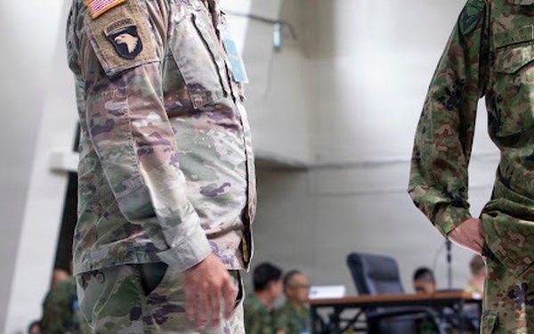 The image shows military personnel from at least two different nations standing in what appears to be a command or operations center. A U.S. Army soldier wearing OCP (Operational Camouflage Pattern) with a 101st Airborne Division patch and American flag is visible alongside personnel in a different camouflage pattern consistent with Japan Ground Self-Defense Force (JGSDF) uniforms. The setting suggests a joint military exercise or coordination meeting.