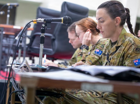 Two female military personnel in Australian Army camouflage uniforms are seated at a desk, appearing to be engaged in a briefing or administrative task. Both wear the Australian national flag patch on their right shoulders, identifying them as members of the Australian Defence Force. A desktop microphone on a stand is positioned in the foreground, suggesting a formal meeting or conference setting.