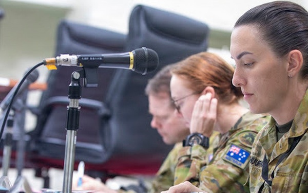Two female military personnel in Australian Army camouflage uniforms are seated at a desk, appearing to be engaged in a briefing or administrative task. Both wear the Australian national flag patch on their right shoulders, identifying them as members of the Australian Defence Force. A desktop microphone on a stand is positioned in the foreground, suggesting a formal meeting or conference setting.