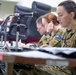 Two female military personnel in Australian Army camouflage uniforms are seated at a desk, appearing to be engaged in a briefing or administrative task. Both wear the Australian national flag patch on their right shoulders, identifying them as members of the Australian Defence Force. A desktop microphone on a stand is positioned in the foreground, suggesting a formal meeting or conference setting.