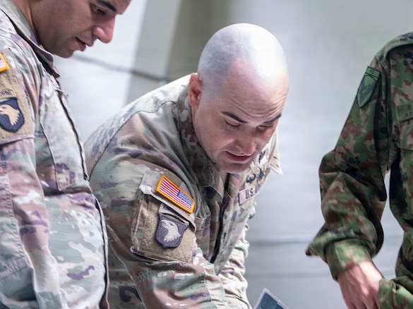 The image shows two U.S. Army soldiers in MultiCam (OCP) uniforms alongside a third individual in a different camouflage pattern, likely a foreign military uniform. The U.S. soldiers are wearing 101st Airborne Division patches and U.S. flag patches on their right sleeves, suggesting a joint or multinational military activity. All three individuals appear to be looking downward, possibly reviewing documents or equipment.