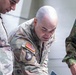 The image shows two U.S. Army soldiers in MultiCam (OCP) uniforms alongside a third individual in a different camouflage pattern, likely a foreign military uniform. The U.S. soldiers are wearing 101st Airborne Division patches and U.S. flag patches on their right sleeves, suggesting a joint or multinational military activity. All three individuals appear to be looking downward, possibly reviewing documents or equipment.
