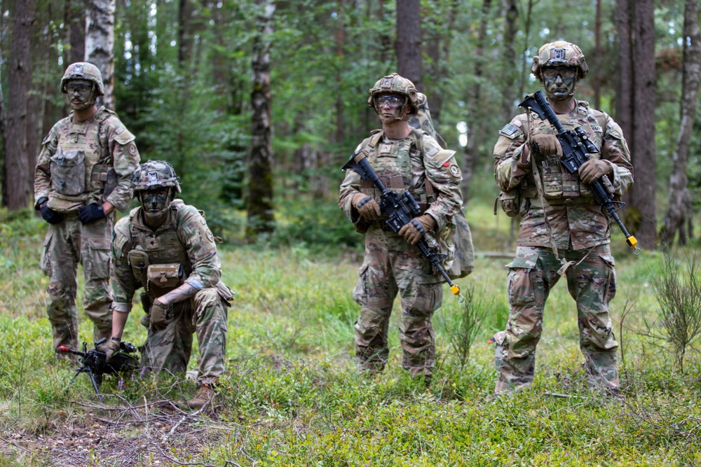 Four military personnel in full combat gear are positioned in a forested area, wearing MultiCam/OCP camouflage uniforms with face paint applied. Three soldiers are standing while one is kneeling, appearing to handle a small drone on the ground. The setting and equipment suggest a training exercise rather than active combat.