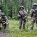 Four military personnel in full combat gear are positioned in a forested area, wearing MultiCam/OCP camouflage uniforms with face paint applied. Three soldiers are standing while one is kneeling, appearing to handle a small drone on the ground. The setting and equipment suggest a training exercise rather than active combat.