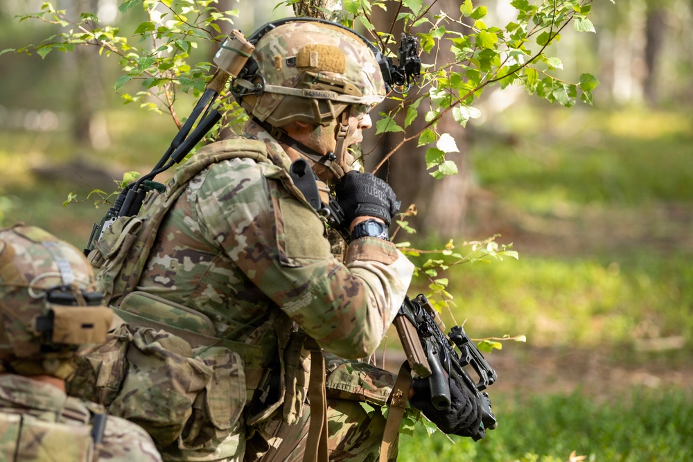 A soldier in full multicam/OCP camouflage kit is positioned in a wooded area, appearing to communicate via radio or hand-held device while holding a weapon. A second soldier in similar gear is partially visible in the foreground left. The setting suggests a field training exercise or operational patrol in a temperate forest environment.
