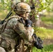A soldier in full multicam/OCP camouflage kit is positioned in a wooded area, appearing to communicate via radio or hand-held device while holding a weapon. A second soldier in similar gear is partially visible in the foreground left. The setting suggests a field training exercise or operational patrol in a temperate forest environment.