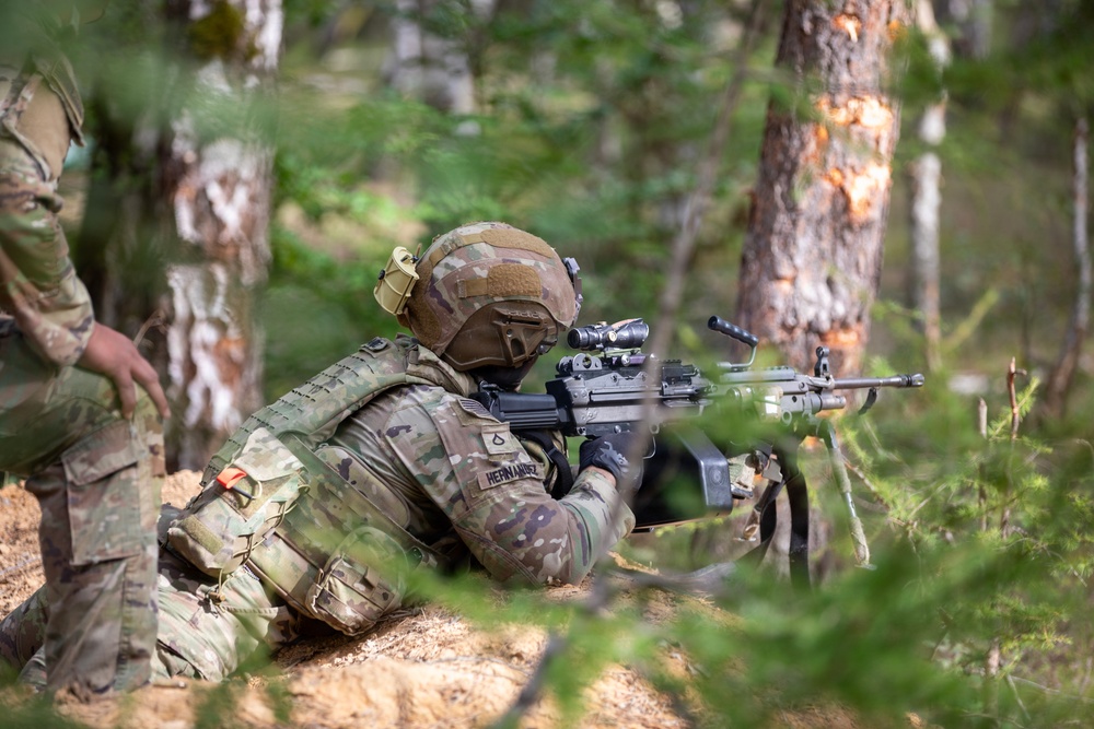 A U.S. Army soldier identified by name tape as 'Hernandez' is in a prone firing position in a forested environment, aiming a weapon equipped with an optic scope and vertical foregrip. The soldier is wearing MultiCam (OCP) camouflage uniform, plate carrier vest, tactical helmet with accessories, and black gloves. A second soldier in similar uniform is partially visible on the left side of the frame.
