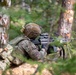 A U.S. Army soldier identified by name tape as 'Hernandez' is in a prone firing position in a forested environment, aiming a weapon equipped with an optic scope and vertical foregrip. The soldier is wearing MultiCam (OCP) camouflage uniform, plate carrier vest, tactical helmet with accessories, and black gloves. A second soldier in similar uniform is partially visible on the left side of the frame.