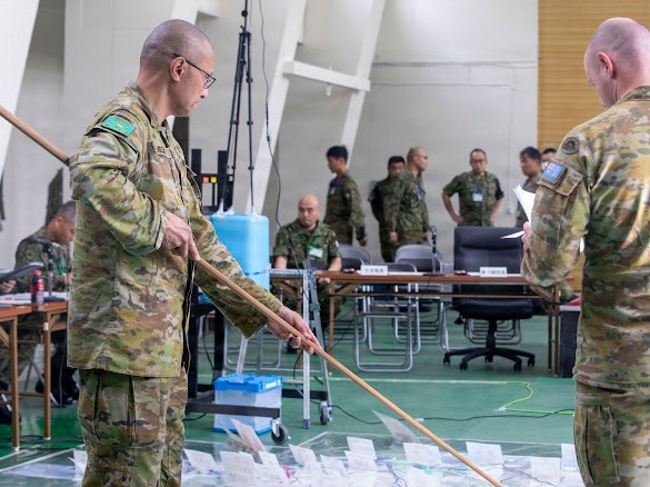 Military personnel in camouflage uniforms are engaged in a command post or tactical planning exercise, with two foreground figures using a long pointer/stick to indicate positions on a floor map. The setting appears to be an indoor operations room with tables, documents, and multiple personnel from what appear to be different national militaries collaborating. Visible national flag patches suggest Australian Defence Force participation alongside Asian military counterparts, likely Japanese Self-Defense Forces.