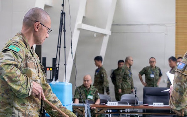Military personnel in camouflage uniforms are engaged in a command post or tactical planning exercise, with two foreground figures using a long pointer/stick to indicate positions on a floor map. The setting appears to be an indoor operations room with tables, documents, and multiple personnel from what appear to be different national militaries collaborating. Visible national flag patches suggest Australian Defence Force participation alongside Asian military counterparts, likely Japanese Self-Defense Forces.