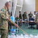 Military personnel in camouflage uniforms are engaged in a command post or tactical planning exercise, with two foreground figures using a long pointer/stick to indicate positions on a floor map. The setting appears to be an indoor operations room with tables, documents, and multiple personnel from what appear to be different national militaries collaborating. Visible national flag patches suggest Australian Defence Force participation alongside Asian military counterparts, likely Japanese Self-Defense Forces.