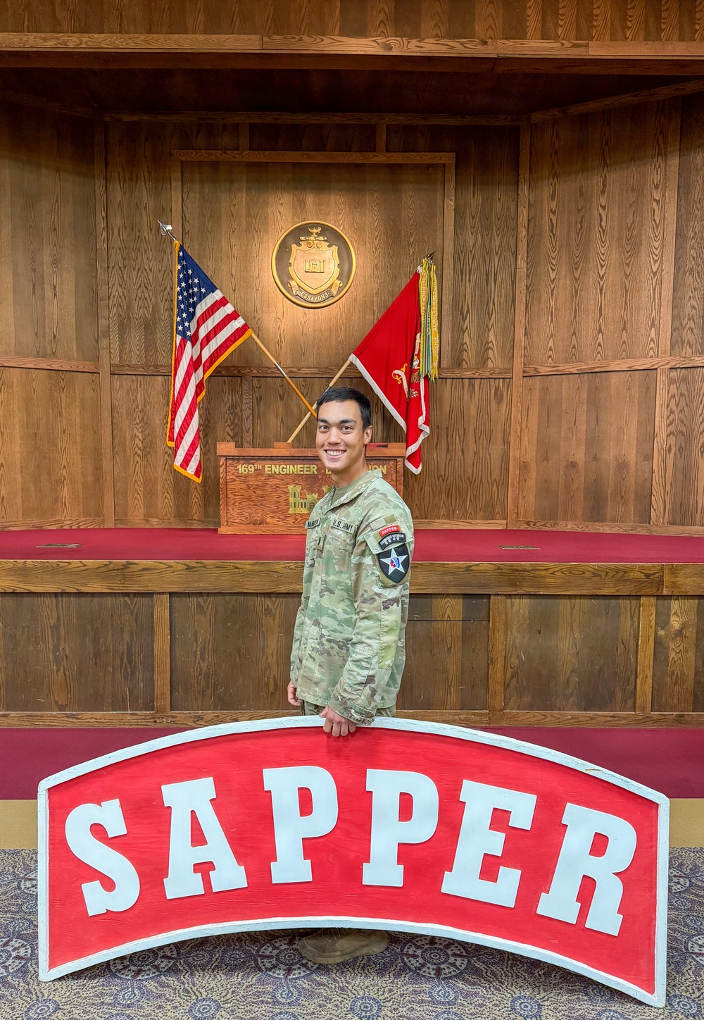 A U.S. Army soldier in OCP (Operational Camouflage Pattern) uniform stands smiling in front of a large red 'SAPPER' tab sign. Behind him is a formal military display area featuring crossed U.S. and unit flags, a wooden podium reading '169th Engineer Battalion,' and a unit crest mounted on a wood-paneled wall. The soldier wears a 2nd Infantry Division patch and a 'SAPPER' tab on his left shoulder.