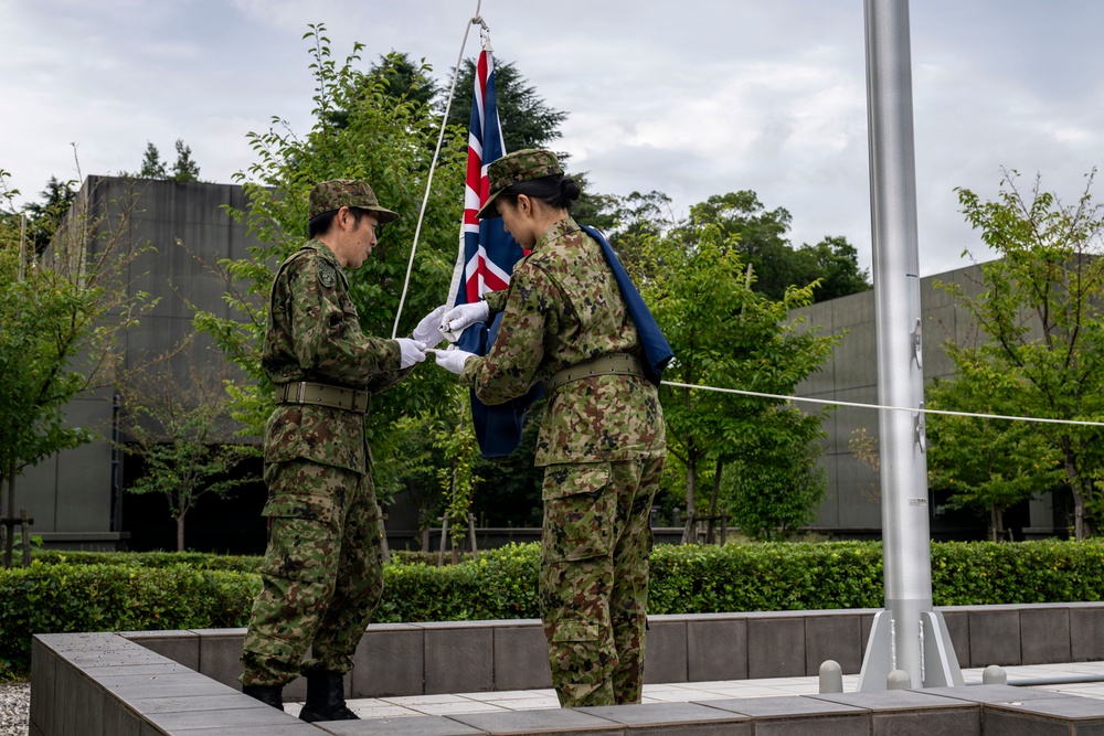 Two military personnel in green camouflage uniforms are handling a flag at a flagpole on an elevated platform. The flag being prepared appears to be the Australian or New Zealand flag (featuring the Union Jack design). The setting appears to be a formal military or government compound with manicured hedges and modern buildings in the background.