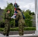 Two military personnel in green camouflage uniforms are handling a flag at a flagpole on an elevated platform. The flag being prepared appears to be the Australian or New Zealand flag (featuring the Union Jack design). The setting appears to be a formal military or government compound with manicured hedges and modern buildings in the background.