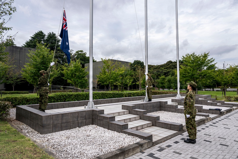 Three military personnel in camouflage uniforms are conducting a flag-raising ceremony at a formal outdoor plaza. The Australian flag is being raised on one of three flagpoles at what appears to be a military installation or memorial site. The setting features a structured stone plaza with steps, white gravel, and manicured greenery in the background.