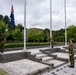 Three military personnel in camouflage uniforms are conducting a flag-raising ceremony at a formal outdoor plaza. The Australian flag is being raised on one of three flagpoles at what appears to be a military installation or memorial site. The setting features a structured stone plaza with steps, white gravel, and manicured greenery in the background.