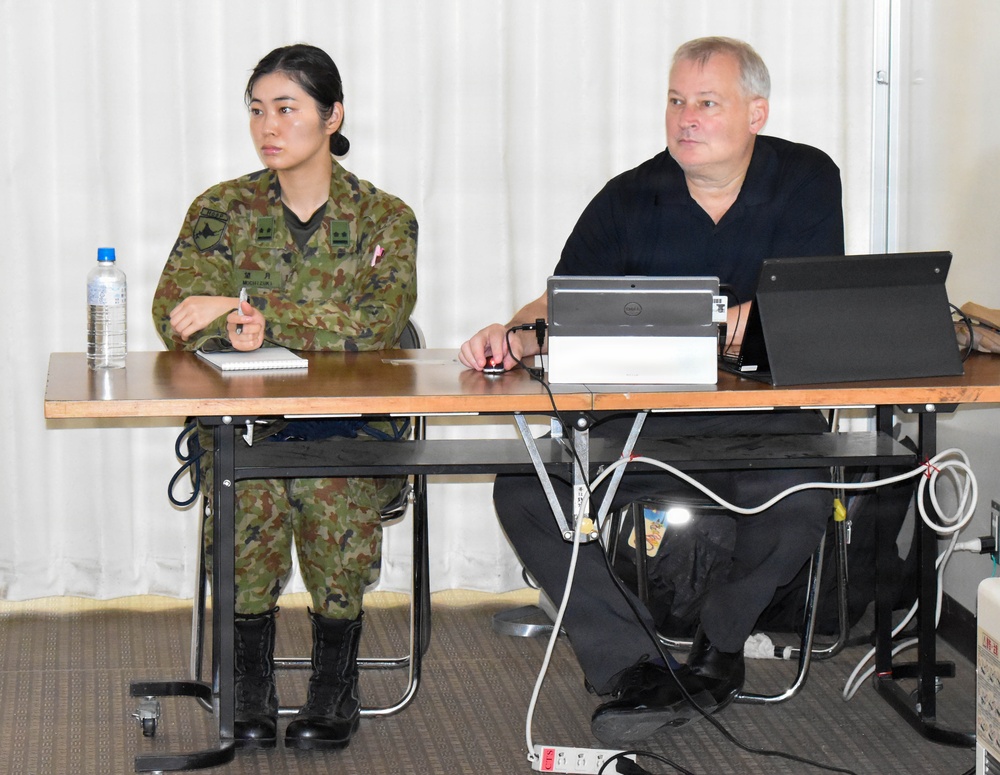 Two individuals are seated at a table in what appears to be a briefing or meeting room. One person is wearing Japanese Ground Self-Defense Force (JGSDF) camouflage uniform, and the other is a civilian in a black shirt. A Dell laptop and an additional monitor are set up on the table, suggesting a presentation or working session.